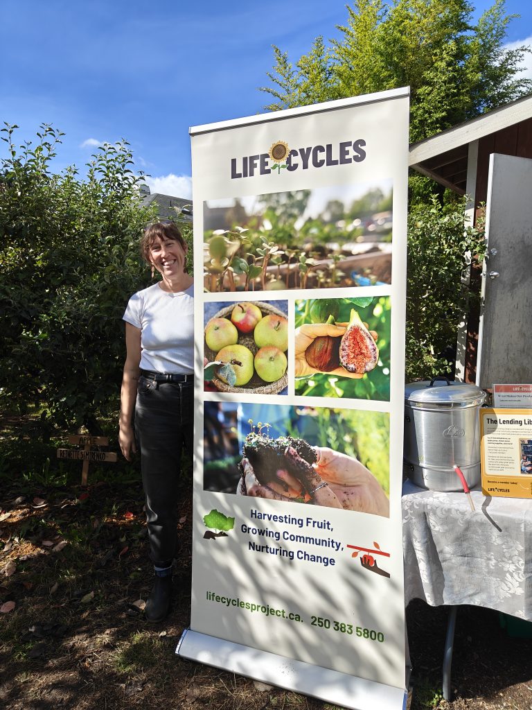 A LifeCycles staff member smiling beside a tall LifeCycles banner featuring photos of harvested fruit and the tagline "Harvesting Fruit, Growing Community, Nurturing Change".