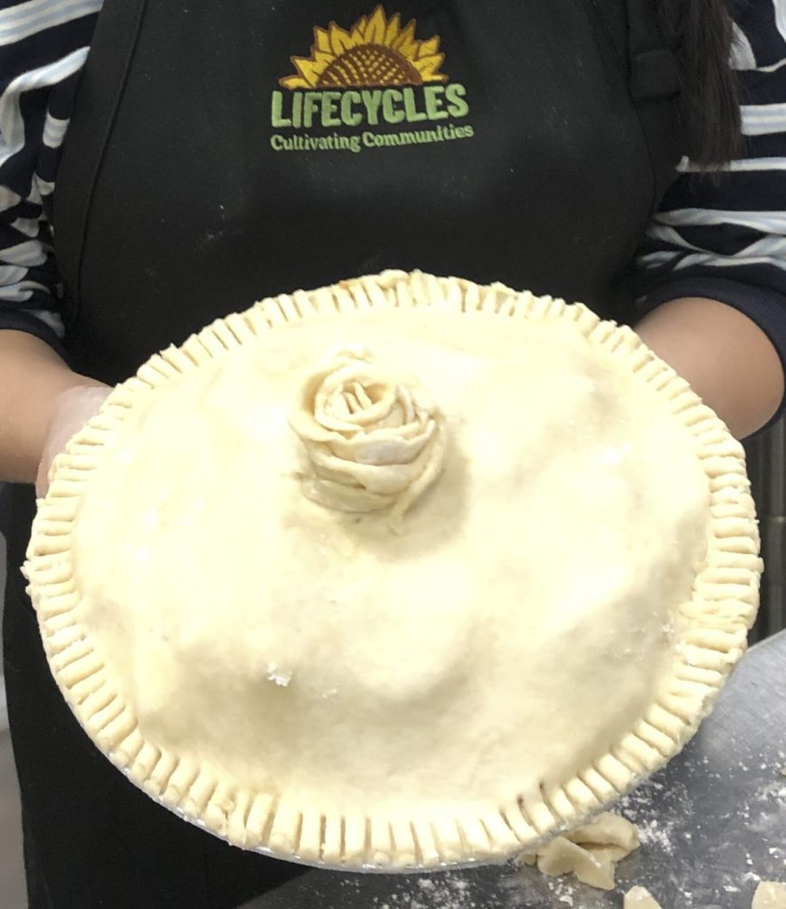 A person wearing a LifeCycles apron holding an unbaked homemade pie topped with a decorative pastry rose.